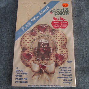 May include: A wood craft kit featuring a gingerbread man with a wreath of hearts. The kit includes 10 wood cut-outs with paste-on patterns. The text on the wreath reads "I Love My Country Kitchen".