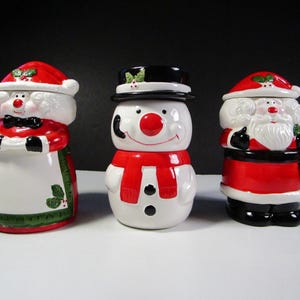 May include: Three ceramic Christmas cookie jars featuring Santa Claus, a snowman, and a woman wearing a red dress and a white apron. The jars are decorated with holly and other festive details.