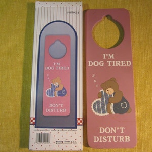 May include: Pink wooden door hanger with a bear holding a heart-shaped pillow. The hanger has a hole at the top for hanging on a door. The text on the hanger reads "I'm Dog Tired" and "Don't Disturb".