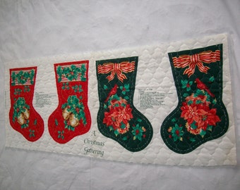 May include: A fabric panel with instructions for making Christmas stockings. The panel features three different designs of stockings with red, green, and gold accents. The text on the panel reads "A Christmas Gathering".