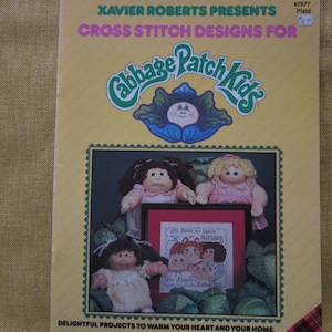 May include: A cross stitch pattern book featuring Cabbage Patch Kids. The cover features a colorful illustration of a Cabbage Patch Kid doll with a green cabbage leaf and a blue flower. The title of the book is "Cabbage Patch Kids" and the subtitle is "Cross Stitch Designs For".