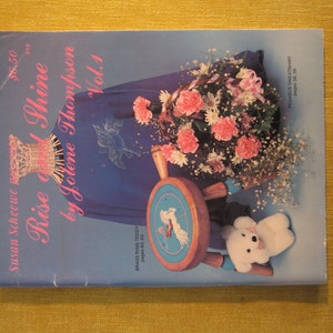 May include: A book cover with the title "Rise and Shine" by Jotene Thompson, Vol. 1. The cover features a blue and white teddy bear, a wooden stool with a blue and white embroidered top, and a bouquet of pink and white flowers.