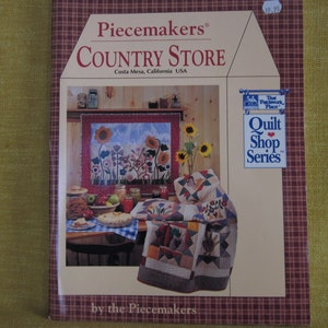 May include: A book cover with a red and white plaid background. The title is "Piecemakers® COUNTRY STORE" with the subtitle "Costa Mesa, California USA". The cover features a quilt shop scene with a quilt hanging on the wall and a quilt folded on a table. The text "Quilt Shop Series" is in a blue box with a red heart.