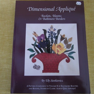 May include: Book cover with a purple background and a colorful illustration of a flower arrangement in a red vase. The title is "Dimensional Appliqué: Baskets, Blooms & Baltimore Borders" by Elly Sienkiewicz. A pattern companion to Volume II of Baltimore Beauties and Beyond, Studies in Classic Album Quilt Appliqué.