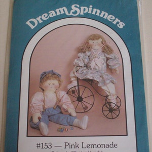 May include: A sewing pattern for a 22-inch doll, featuring a girl doll in a pink and white dress riding a tricycle and a boy doll in blue overalls. The pattern is titled "#153 - Pink Lemonade" and is copyrighted 1988 by Nancy J. Smith and Lynda S. Milligan.