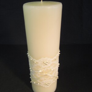 May include: A white pillar candle with a white lace ribbon and pearl accents. The candle is on a black background.