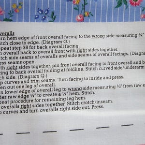 May include: A close-up of a sewing pattern for overalls. The instructions include steps for sewing the side seams, pressing seams open, and turning the hem edge of the front overall facing to the wrong side measuring 1/2 inch.