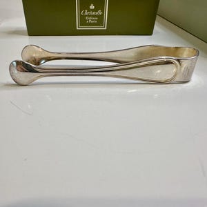 May include: Silver-toned sugar tongs with rounded ends and a curved handle. The tongs are resting on a white surface, with a green box in the background. The box has a white label with the text "Christofle Orfèvre à Paris".