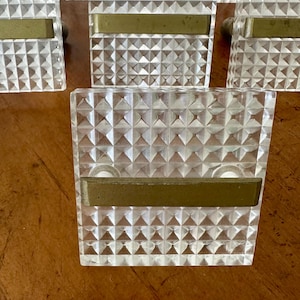 May include: Clear, square cabinet knobs with a diamond-patterned surface and a brass-colored bar. The knobs are arranged on a wooden surface. These decorative knobs are suitable for furniture.