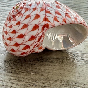 May include: A decorative, shell-shaped ceramic bowl. The bowl is white with a red leaf-like pattern covering the exterior. The interior is a glossy, reflective white. The bowl sits on a light gray wooden surface.