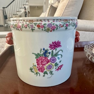 May include: A white ceramic container with a floral design. The container has a band of flowers around the rim and a bouquet of pink, purple, and blue flowers on the side. It has two red handles.