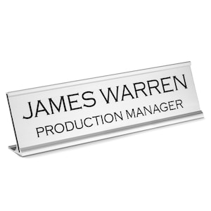 K&ouml;nnte beinhalten: Silberne Schreibtisch-Namensschilder mit schwarzem Text, der "JAMES WARREN PRODUCTION MANAGER" lautet.