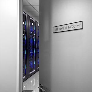 K&ouml;nnte beinhalten: Ein Serverraum mit einem Schild mit der Aufschrift "SERVER ROOM" an der T&uuml;r. Der Raum ist gef&uuml;llt mit schwarzen Serverracks mit blauen Lichtern.