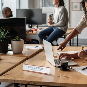 K&ouml;nnte beinhalten: Ein h&ouml;lzerner Schreibtisch-Namensschild mit dem Text "STELLA KEPNES SALES MANAGER" in blauer Schrift. Das Namensschild befindet sich auf einem Holztisch mit einem Laptop, einer Kaffeetasse und einem Notizbuch.