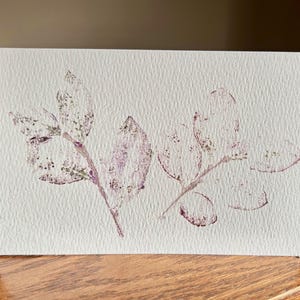 May include: A rectangular white card featuring a botanical print of two leafy branches. The leaves are rendered in a textured, speckled purple and muted green ink, creating a stamped appearance on the thick, slightly rough paper surface.