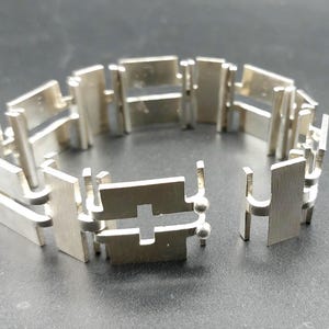 May include: A silver-colored bracelet with a geometric design. The bracelet is composed of rectangular and cross-shaped links, connected by small, rounded elements. The links are flat and have a brushed metal finish. The bracelet is displayed on a dark surface.