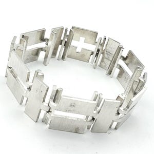 May include: A silver bracelet featuring a geometric design. The bracelet is made up of rectangular and cross-shaped links, giving it a modern, industrial look. The links are joined to create a flexible band.
