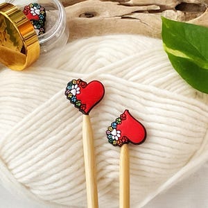 May include: Two wooden knitting needles with red heart-shaped charms. The charms have a white and colorful floral design with paw prints.