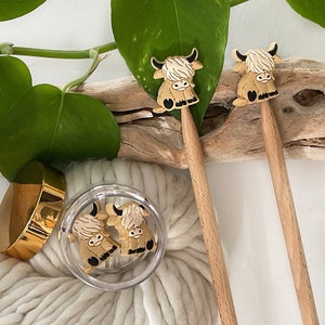 May include: Two wooden knitting needles with brown Highland cow toppers. The needles are resting on a piece of driftwood with white yarn and a jar of additional cow toppers.
