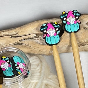 Puede incluir: Dos agujas de tejer de madera con adornos de gnomos. Los adornos son de color rosa y blanco con un sombrero turquesa y dicen "Hello Fall". Un frasco lleno de adornos similares está en el fondo.
