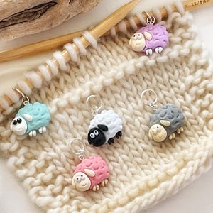 May include: Five colorful sheep stitch markers on a beige knitted fabric. The markers are made of polymer clay and have different colored faces and bodies. The markers are attached to a wooden knitting needle.