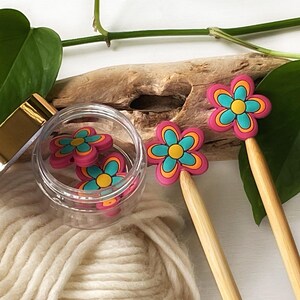May include: A set of colourful flower-shaped knitting needle tips in a glass jar with a gold lid. The tips are pink, blue, and yellow. The tips are on a wooden surface with a ball of yarn.