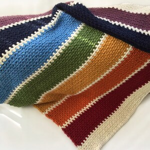 May include: A rainbow striped crochet blanket with a white border. The blanket is folded in half and the colors are purple, navy blue, light blue, green, orange, red, and burgundy.