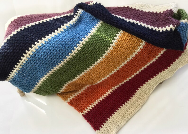 May include: A rainbow striped crochet blanket with a white border. The blanket is folded in half and the colors are purple, navy blue, light blue, green, orange, red, and burgundy.
