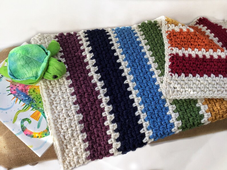 May include: A multi-colored crocheted blanket with stripes of white, purple, navy blue, light blue, green, orange, and red. The blanket is made with a textured stitch and has a fringe border.