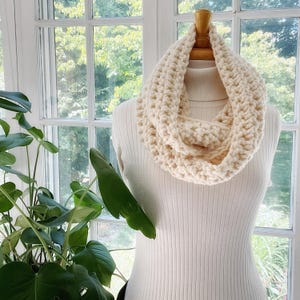 May include: A cream-colored crocheted infinity scarf. The scarf is thick and textured, and it is displayed on a white mannequin.
