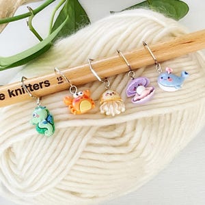 May include: Five colourful sea creature themed stitch markers on a wooden knitting needle. The markers feature a seahorse, crab, jellyfish, clam, and whale.