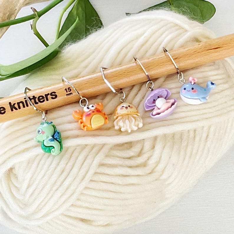 May include: Five colourful sea creature themed stitch markers on a wooden knitting needle. The markers feature a seahorse, crab, jellyfish, clam, and whale.