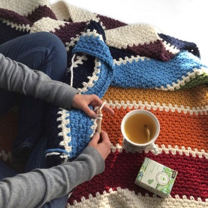 May include: A person crocheting a colorful striped blanket with a cup of tea and a box of herbal tea on a white surface.