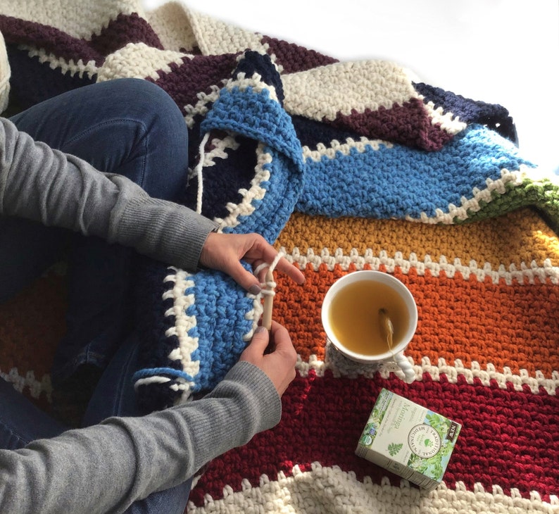 May include: A person crocheting a colorful striped blanket with a cup of tea and a box of herbal tea on a white surface.