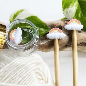 May include: Two wooden knitting needles with rainbow and cloud toppers. The needles are on a wooden surface with white yarn in the background.