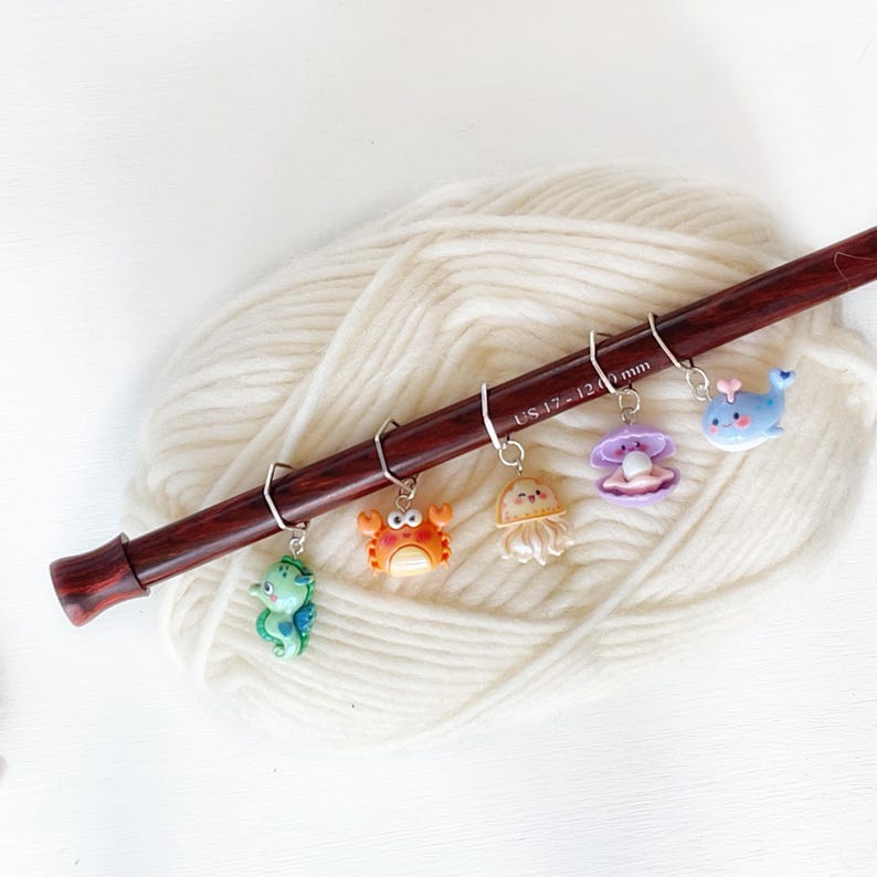 May include: A set of six colourful sea creature stitch markers on a wooden knitting needle. The markers feature a seahorse, crab, jellyfish, clam, whale, and a UK size 12.0 mm knitting needle.