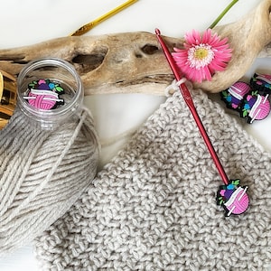 May include: A close-up of a beige crochet project with a red crochet hook and a pink flower. The crochet hook has a pink and white charm attached to it that says "Crochet Fairy"
