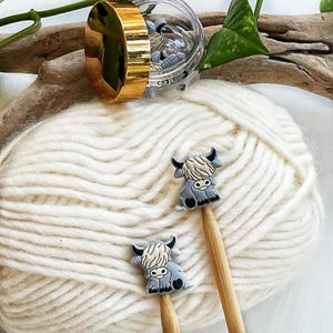 May include: Two small, grey and cream-colored highland cow-shaped charms attached to the ends of wooden knitting needles. The needles are resting on a ball of white yarn, with a clear plastic jar featuring a gold lid containing more charms.