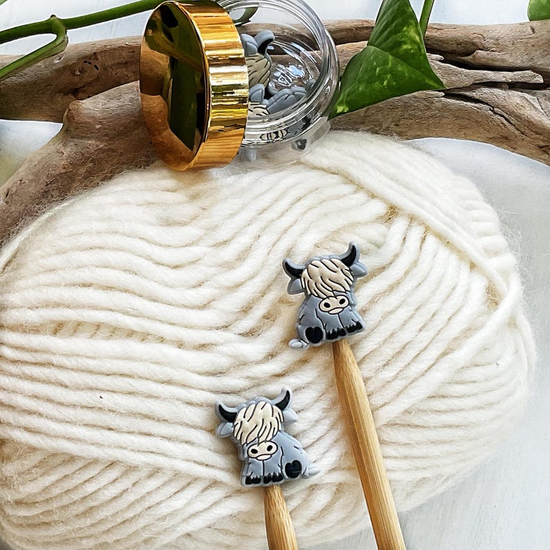 May include: Two small, grey and cream-colored highland cow-shaped charms attached to the ends of wooden knitting needles. The needles are resting on a ball of white yarn, with a clear plastic jar featuring a gold lid containing more charms.