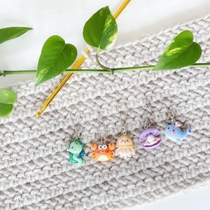 May include: Six colourful crocheting stitch markers shaped like sea creatures, including a crab, a jellyfish, a seahorse, a clam, and a whale. The markers are attached to a beige crocheted fabric.