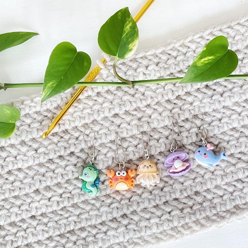 May include: Six colourful crocheting stitch markers shaped like sea creatures, including a crab, a jellyfish, a seahorse, a clam, and a whale. The markers are attached to a beige crocheted fabric.