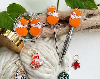 May include: Two metal crochet hooks with orange fox toppers, a clear container holding two orange fox stitch markers, and four additional metal stitch markers featuring a fox, a house, a red maple leaf, and a green mushroom design.
