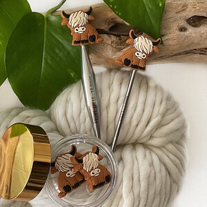 May include: Two knitting needles with brown Highland Cow animal toppers. The needles are made of metal and have a silver finish. The needles are on a white fluffy background. One needle has the text "KnitPro US 15 - 10.00mm" printed on it.