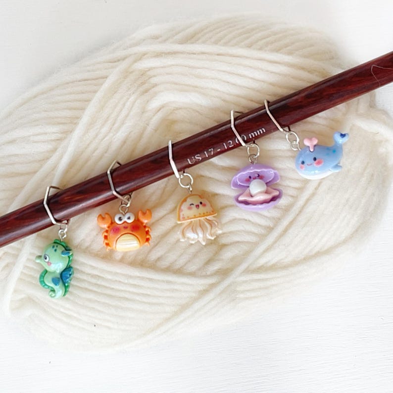 May include: Five colourful sea creature stitch markers on a wooden knitting needle. The markers are a seahorse, crab, jellyfish, clam, and whale. The needle is labelled "US 17 - 12.00 mm".