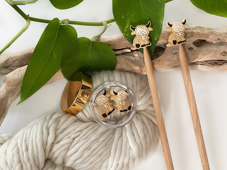 May include: Two wooden knitting needles with brown Highland cow toppers. The needles are resting on a piece of driftwood with white yarn and a jar of additional cow toppers.