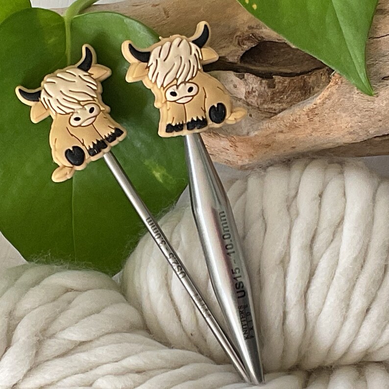 May include: Two silver knitting needles with brown and white Highland Cow animal toppers. The needles are labeled "Knitters Pride US 15 - 10.0 mm" and "US 7 - 4.5 mm".