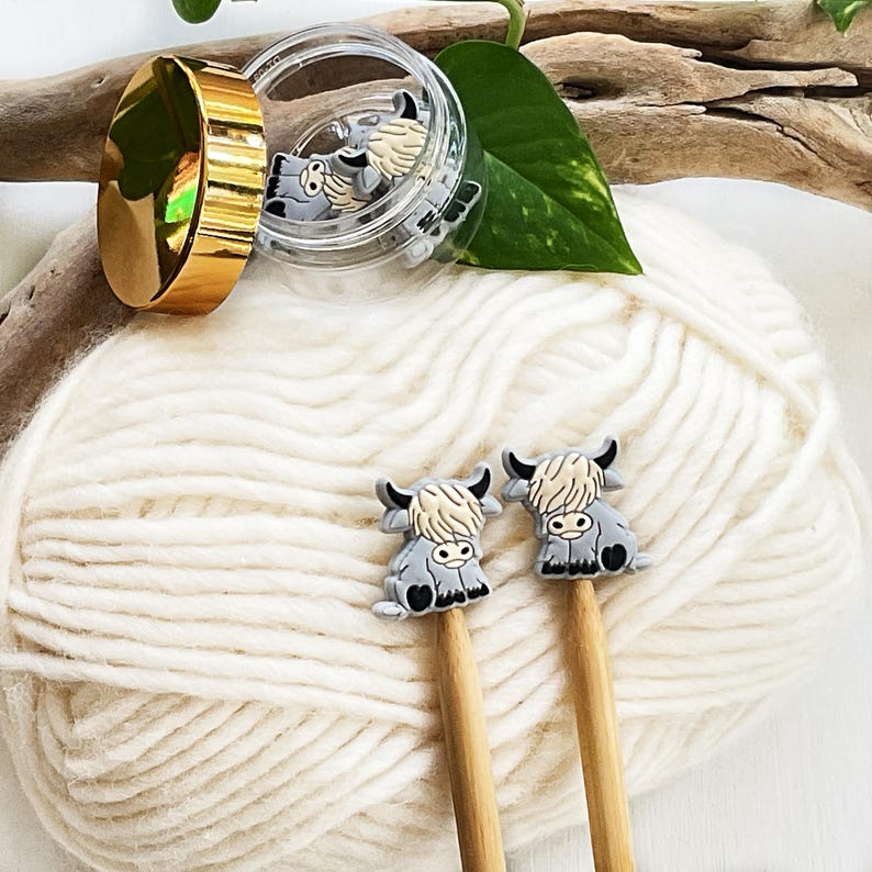 May include: Two small gray and cream-colored highland cow charms attached to the ends of wooden knitting needles. A clear plastic container with a gold lid sits nearby, containing additional identical highland cow charms.