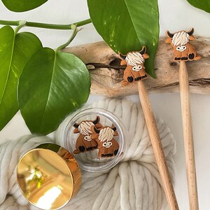 May include: Three brown Highland cow charms on wooden knitting needles. The charms are attached to the needles with a gold ring. The needles are resting on a white fluffy surface.