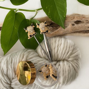 May include: Two knitting needles with Highland Cow animal toppers. The needles are silver and the cow toppers are brown and white. The needles are resting on a white yarn ball.