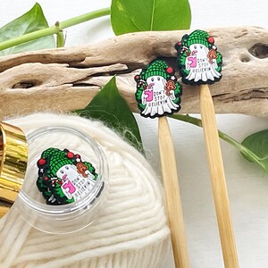 May include: A close-up of three knitting needle toppers with a ghost design. The toppers are white with a green hat and a wreath. The text "DON'T STOP BELIEVIN" is visible. One topper is in a clear container, and two are on wooden needles.